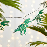 Grüne Dino-Lichterkette mit warmweißen LEDs zwischen Pflanzen und hellem Lichterhintergrund.