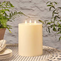 Große cremefarbene LED-Kerze mit drei Flammen auf einem Gartentisch neben grünen Pflanzen.