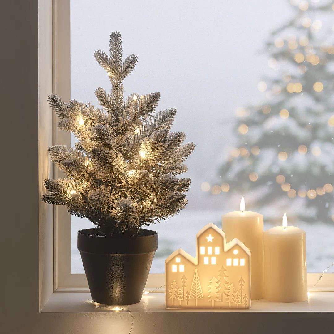 Verschneiter LED-Weihnachtsbaum mit warmweißen Lichtern auf Fensterbank neben Kerzen und Dekohaus
