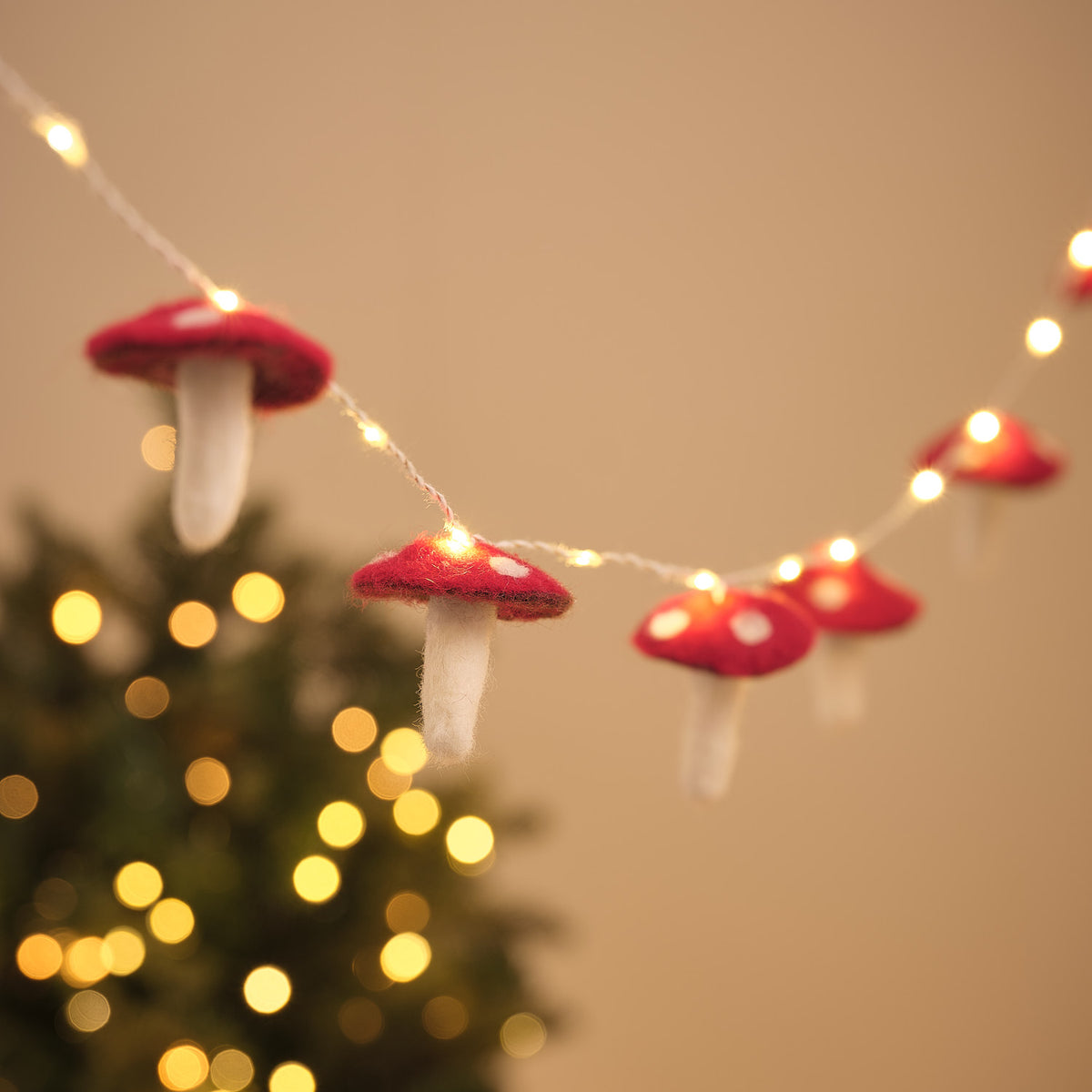 Lichterkette mit Filz-Fliegenpilzen in Rot mit weißen Punkten, warmweiß beleuchtet, vor einem geschmückten Weihnachtsbaum mit Bokeh-Lichtern.