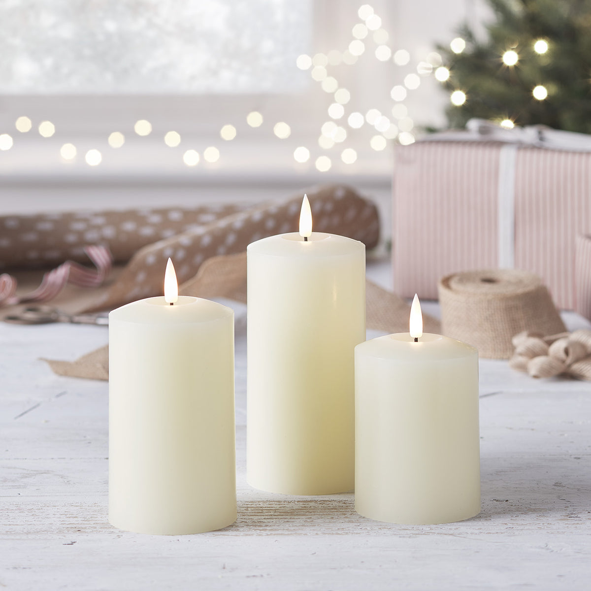 Drei TruGlow LED-Kerzen auf weißem Holz mit Weihnachtsdeko und Geschenk im Hintergrund