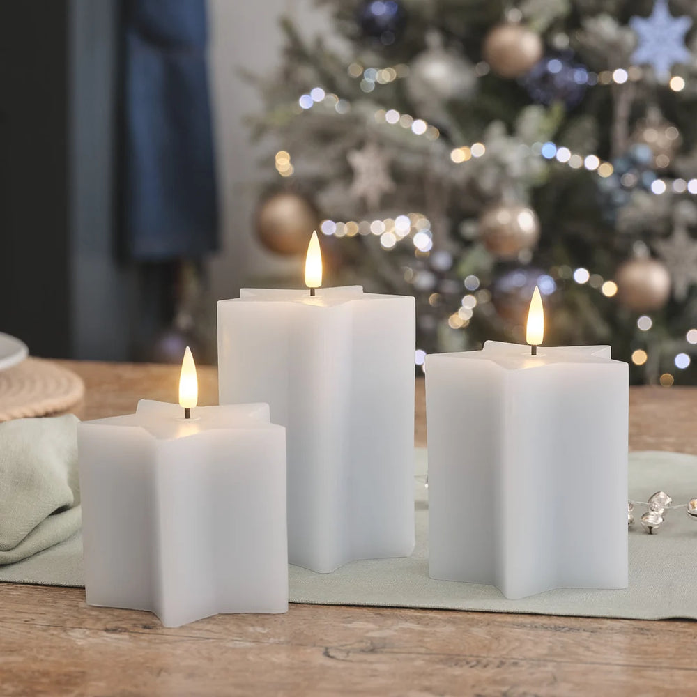 Weiße LED-Kerzen in verschiedenen Höhen mit realistischen Flammen auf Holztisch, Weihnachtsbaum mit bokeh LED-Lichtern im Hintergrund, festliche Dekoration.