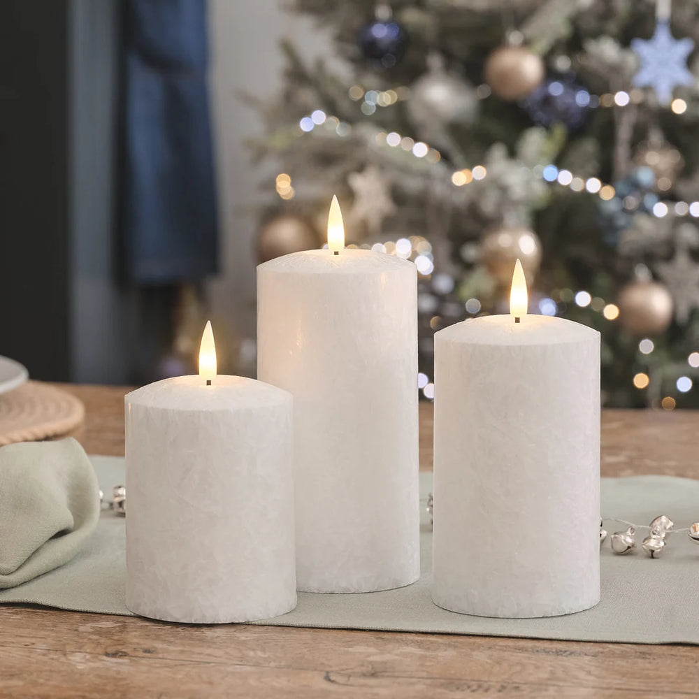 Drei weiße LED-Kerzen in verschiedenen Höhen mit warmweißen LED-Flammen auf Holztisch, beige Leinenserviette, verschwommener Weihnachtsbaum mit Kugeln im Hintergrund.