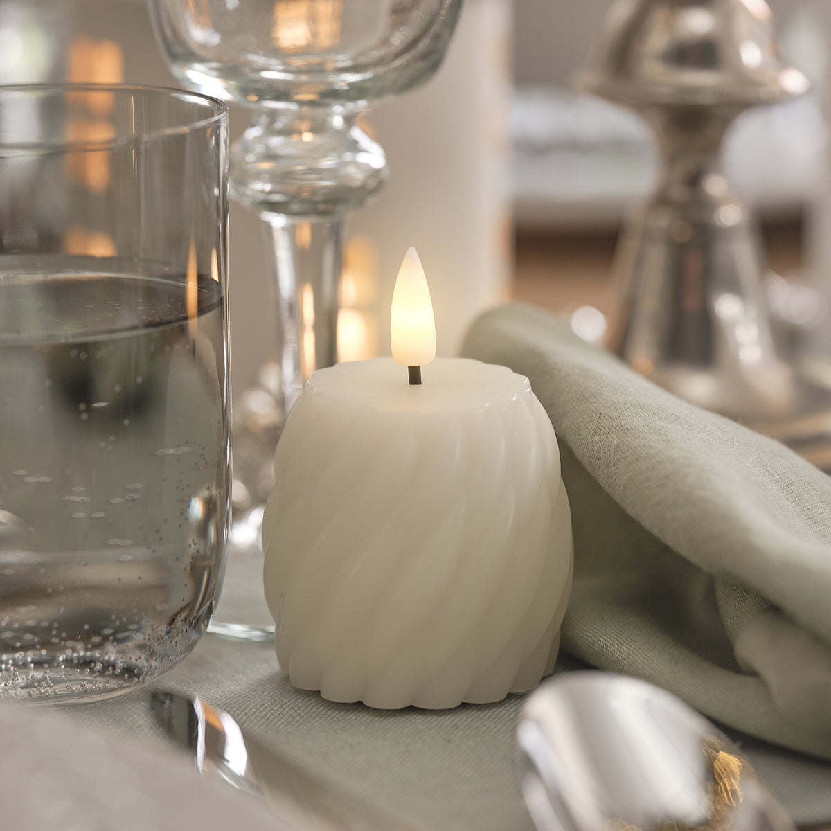 Weiße LED-Kerze mit wellenförmiger Oberfläche und warmweißer LED-Flamme, neben Glas mit Wasser und verschwommenen Leuchtern im Hintergrund.