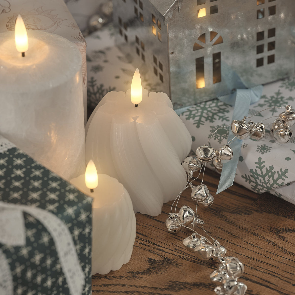 Weiße wellenförmige LED-Kerzen mit warmweißen LED-Flammen, grüne und weiße Leinenservietten mit Schneeflocken-Muster, silberne Weihnachtsglocken auf dunklem Holztisch.