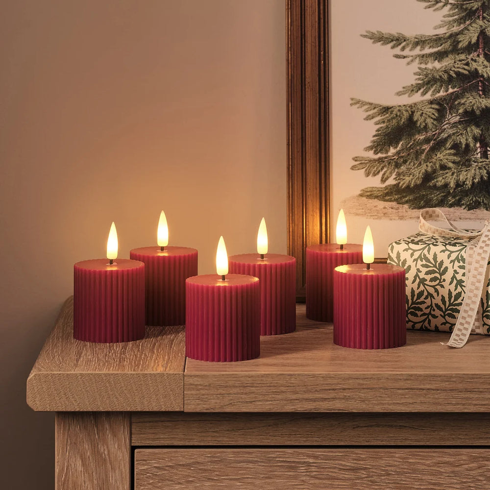 Sechs rote strukturierte LED-Zylinderkerzen mit warmweißen LED-Flammen auf gespaltener Holzoberfläche, beige und dunkles Holz, grüne Girlande mit goldenen Lichtern, dekoratives Leinen-Kissen mit Muster im Hintergrund.