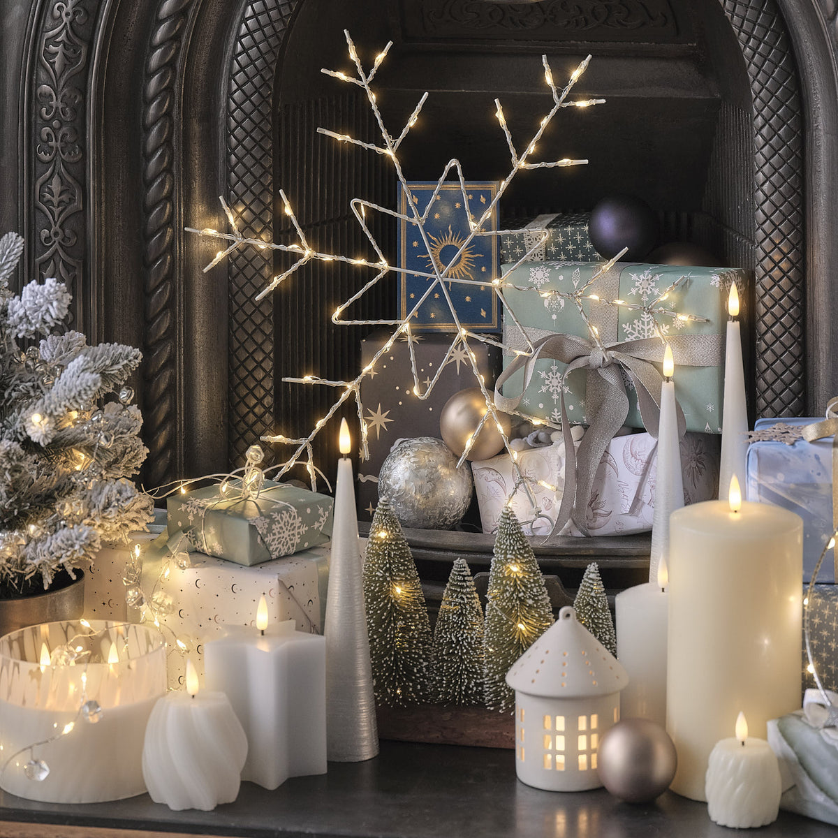 Aufwendige Kaminsims-Dekoration mit weißen LED-Kerzen in Blütenform, cremefarbenen LED-Zylinderkerzen, weißem beleuchteten Weihnachtshaus mit LED-Fenstern, silbernem Ast-Dekorationslicht, grünen schneebedeckten Kegelkerzen-Dekos, silbernen Weihnachtskugeln, Geschenkboxen mit Schneemuster und grauem Band, dunkler Kaminsims mit ornamentalem Muster.