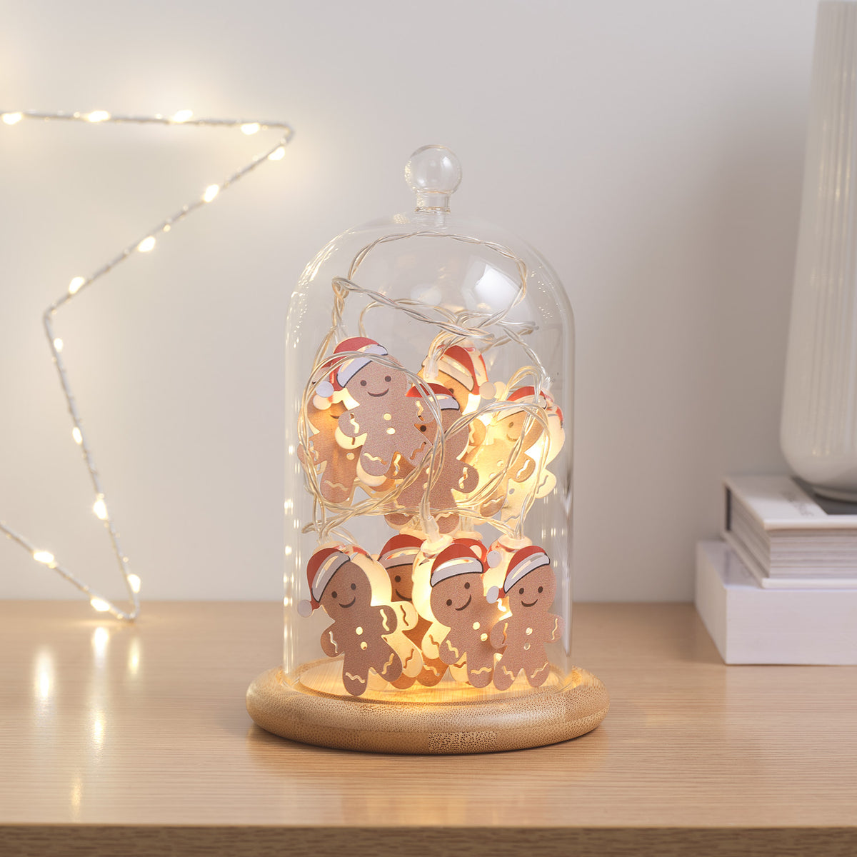 Glasglocke mit Holzteller und leuchtender Lebkuchenmännchen-Lichterkette auf hellem Tisch