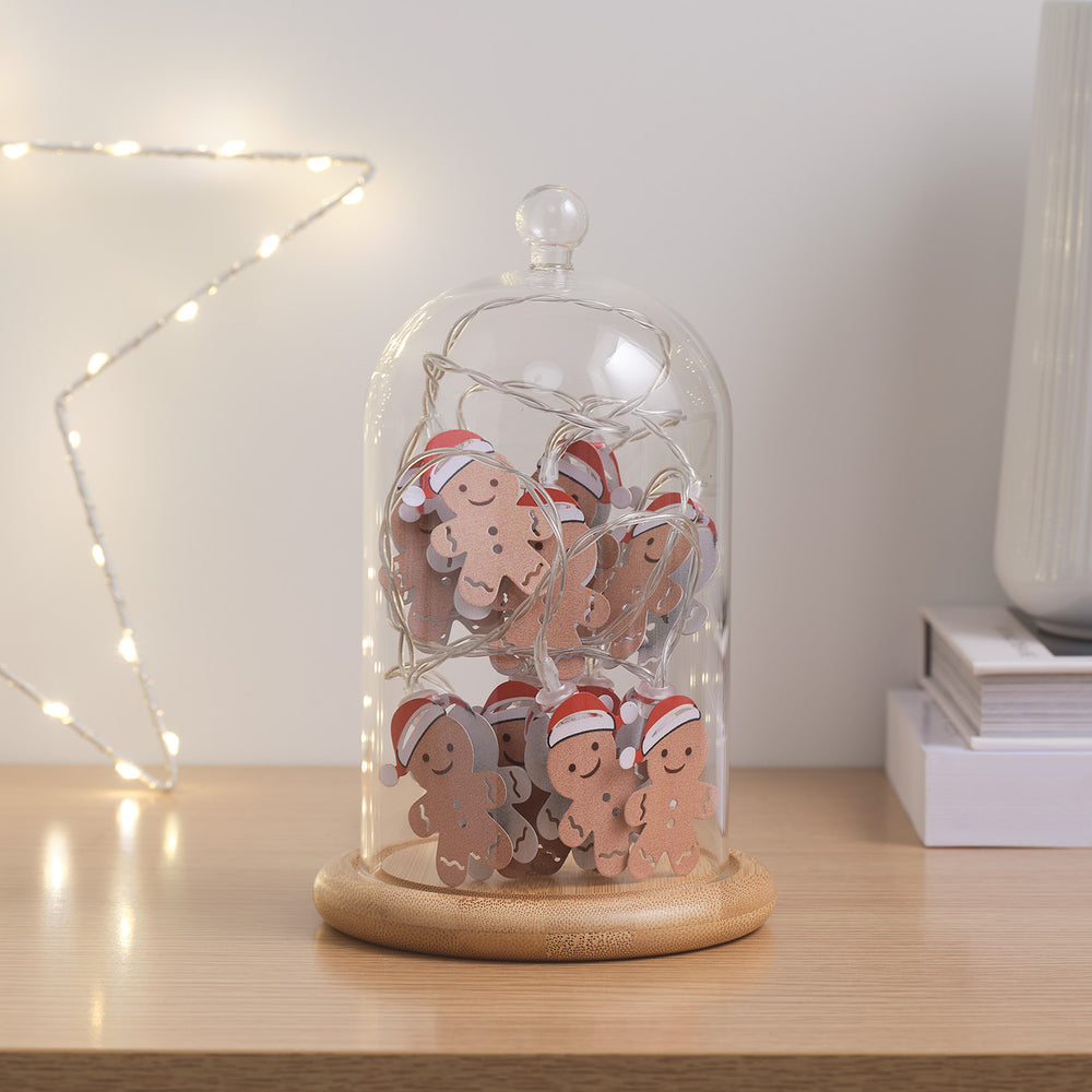 Glasglocke mit Holzteller und unbeleuchteter Lebkuchenmännchen-Lichterkette auf hellem Tisch