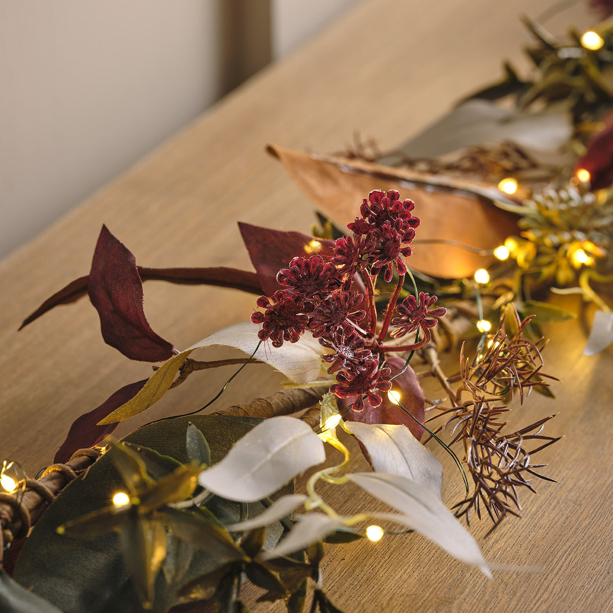Nahaufnahme der herbstlichen Girlande mit roten Blüten, Blättern und warmen LED-Lichtern auf Holztisch
