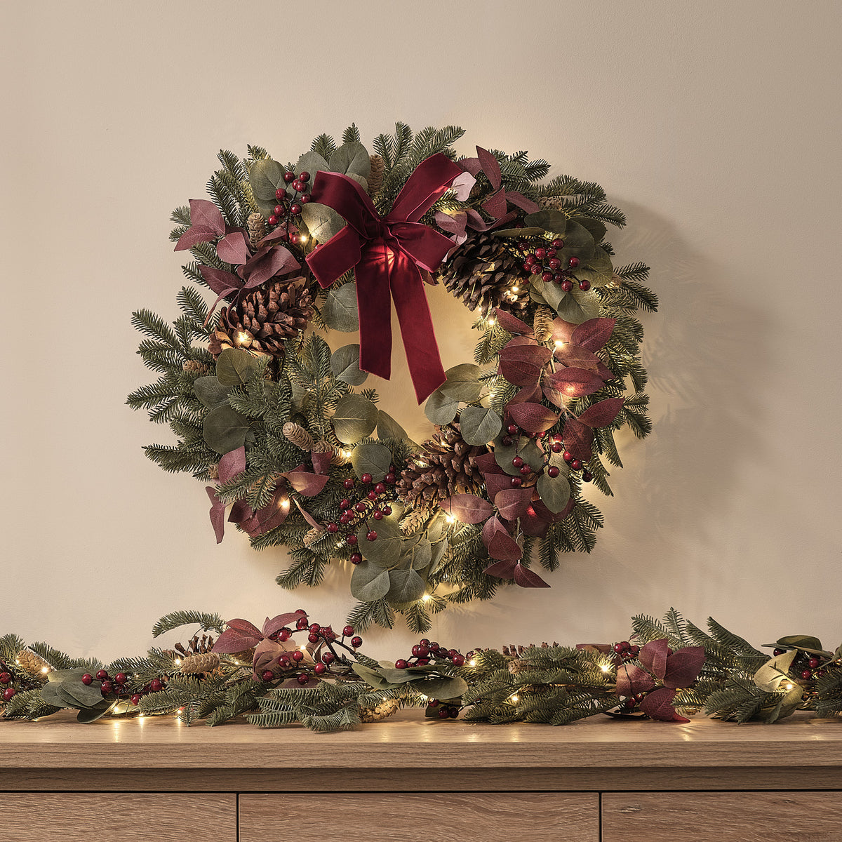 Beleuchteter Weihnachtskranz mit Tannenzapfen, roten Beeren und Schleife an Wand