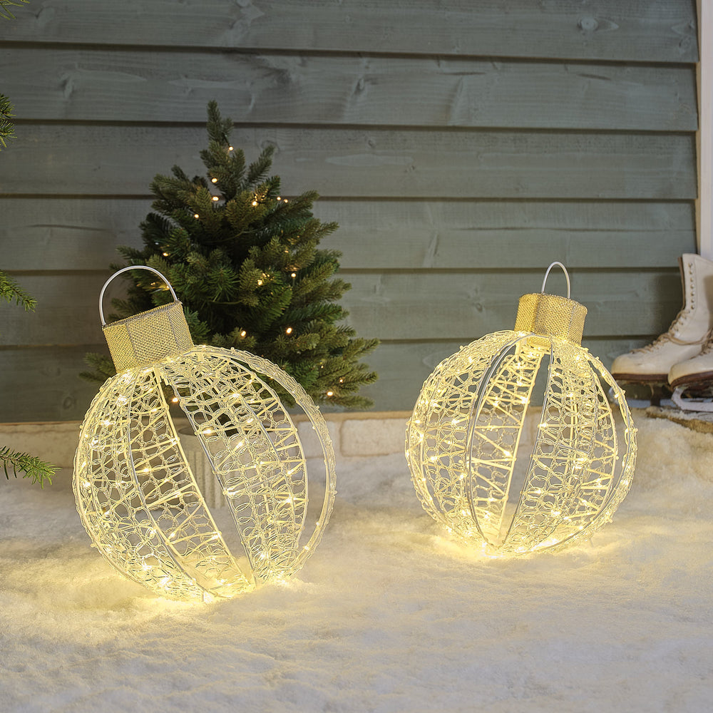 Zwei warmweiße LED-Weihnachtskugeln auf weißem Gestell im Schnee vor grünem Holzhaus