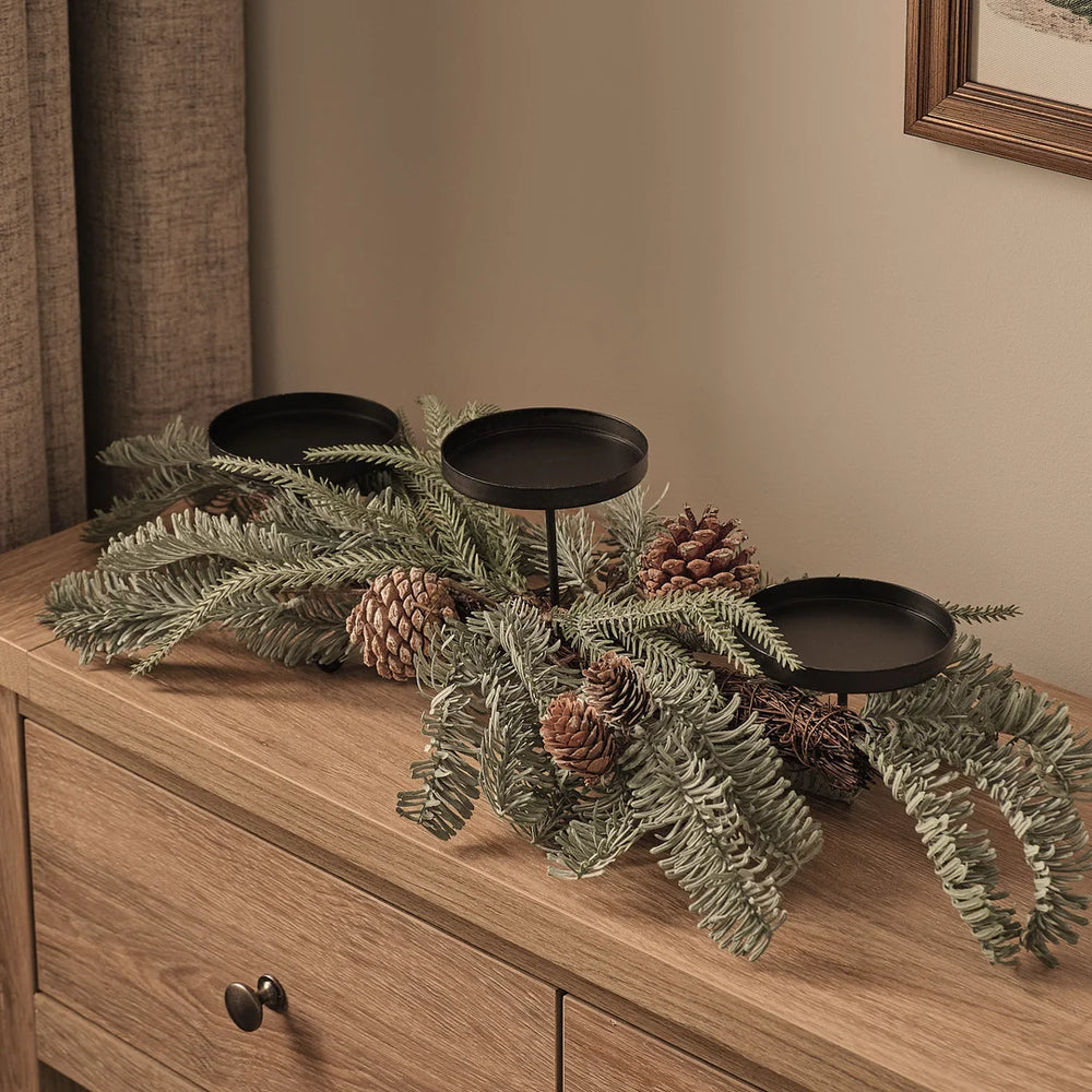 Elegantes Adventsgesteck mit drei schwarzen Kerzenhaltern auf natürlichem Holztisch, dekoriert mit grünem Fichtengrün, braunen Pinienzapfen und natürlichen Waldakzenten – minimalistisches Weihnachtsarrangement für edle Tischdekoration und festliche Wohnzimmer-Displays.
