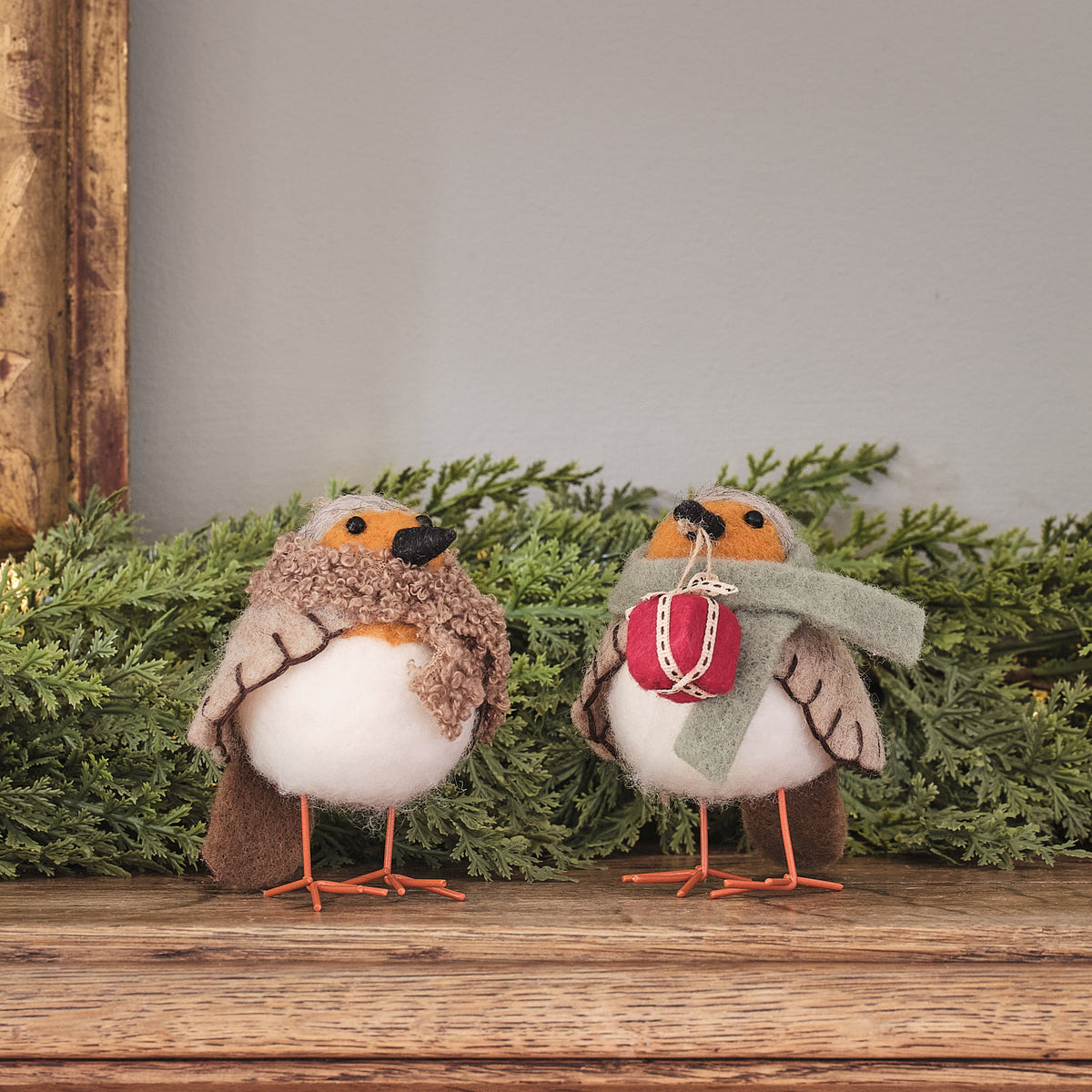 Zwei cremefarbene Vogel-Dekofiguren mit roten gestrickten Weihnachtsmützen und burgundy Schleife, stehend auf grünem Moosuntergrund mit Holzstamm-Hintergrund – niedliche Weihnachtsfiguren für Tischdekoration, Kaminsims und festliche Wohnzimmer-Arrangements mit warmem Charme.