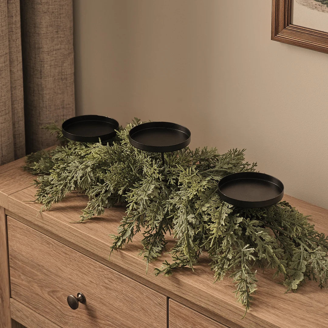 Elegantes lineares Adventsgesteck mit drei schwarzen runden Kerzenhaltern auf natürlichem Holzkommode, dekoriert mit üppigem grünem Fichtengrün in fließender Anordnung und natürlichem Waldcharakter – minimalistische Weihnachtsdekoration für skandinavische Tischlooks und moderne Wohnzimmer-Arrangements.