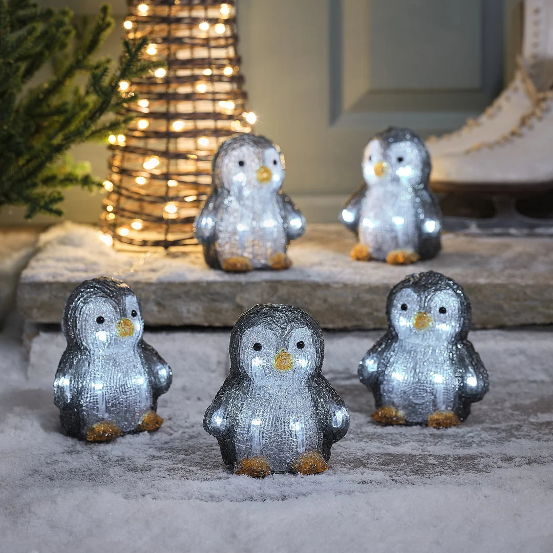 Gruppe von glitzernden grau-weißen Pinguin-Dekofiguren mit Schneeeffekt-Oberflächenfinish und goldenen Details, arrangiert auf grauem Stein-Untergrund vor unscharfem grünem Hintergrund – liebevolle Pinguin-Familie für klassische Weihnachtsarrangements und festliche Außendekoration.