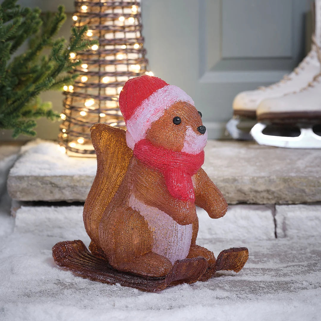 Niedliche glitzernde braune Eichhörnchen-Dekofigur mit roter Weihnachtsmütze und rotem Hals-Accessoire, arrangiert auf Schneeuntergrund vor winterlichem grünem Hintergrund – liebevolle Waldtier-Figur für klassische Weihnachtsarrangements und festliche Außendekoration mit wärmendem Charakter und strukturiertem Glitter-Finish.
