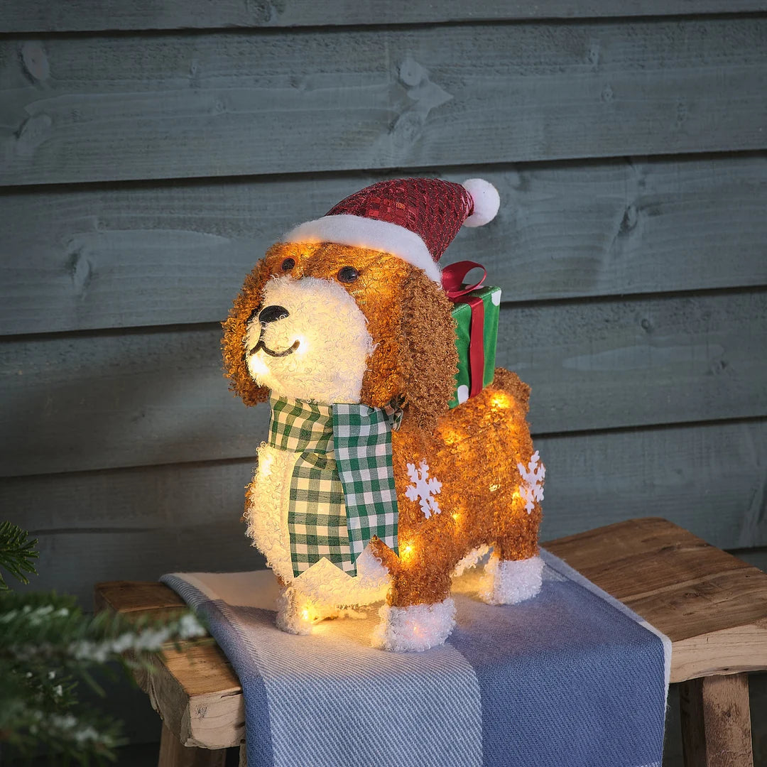 Charmante beleuchtete braun-goldene Teddybär-Dekofigur mit warmweißer LED-Beleuchtung, roter gestrickter Weihnachtsmütze, kariertem grün-weißem Schal und strukturierter Oberfläche, positioniert auf blauem Holzuntergrund mit dunklem Hintergrund – festliche LED-Plüschfigur für Außendekoration, Garten-Arrangements und weihnachtliche Hausdekorationen mit warmem Glühen.