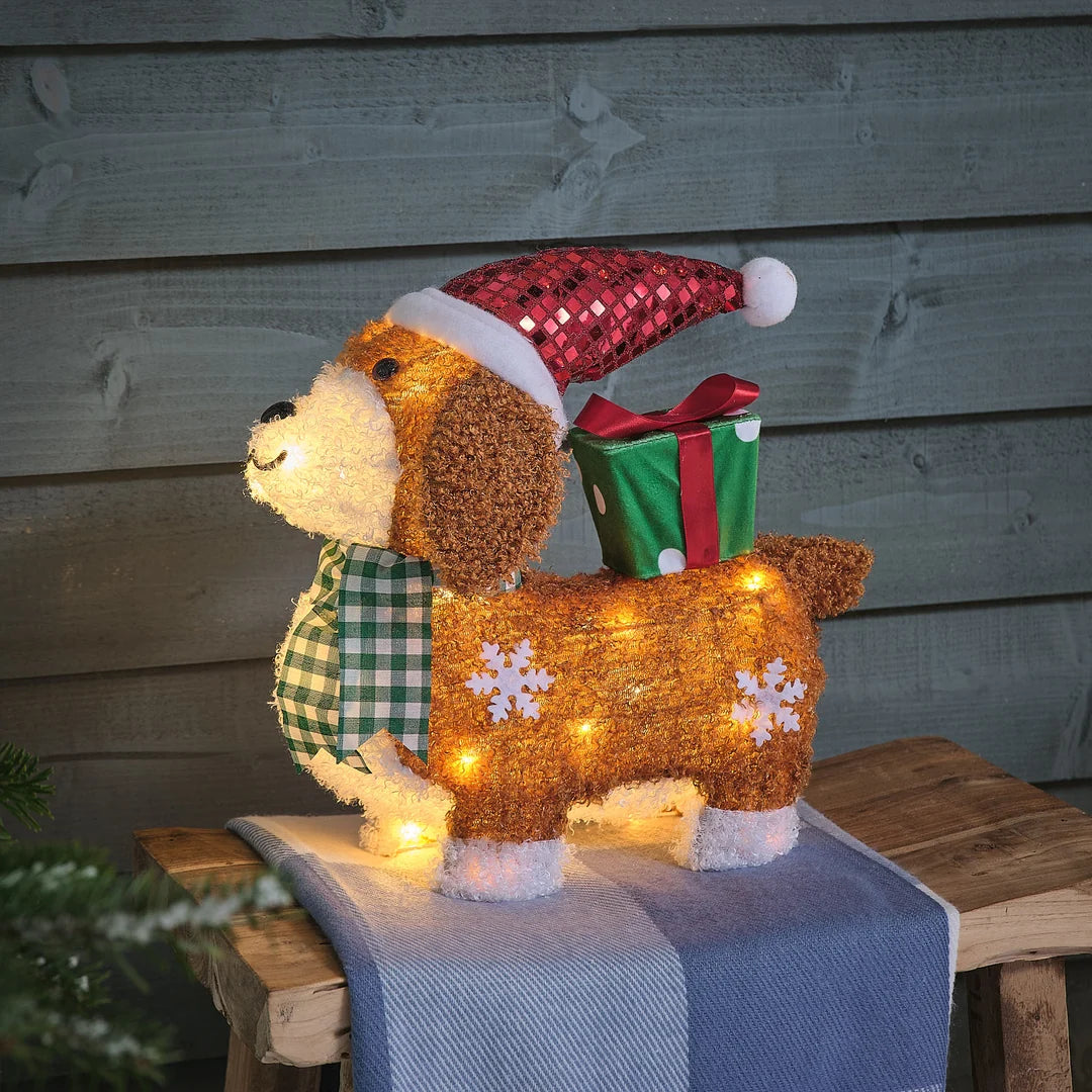 Niedliche beleuchtete hellbraune Teddybär-Dekofigur mit warmweißem LED-Glühen, roter Weihnachtsmütze und kariertem Schal-Accessoire, arrangiert auf blauem Holzblock mit grünem Grünflächen-Hintergrund – liebevolle LED-Plüschfigur für klassische Weihnachtsarrangements und festliche Außendekoration mit atmosphärischer Beleuchtung.