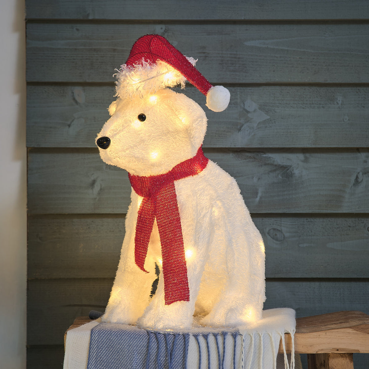Niedliche cremefarbene Plüsch-Affenfigur mit roter gestrickter Weihnachtsmütze und rotem Hals-Accessoire, arrangiert auf blauem Holzblock mit dunklem grauem Hintergrund – liebevolle Tierfigur für klassische Weihnachtsarrangements und festliche Außendekoration mit warmem Weihnachtscharme.