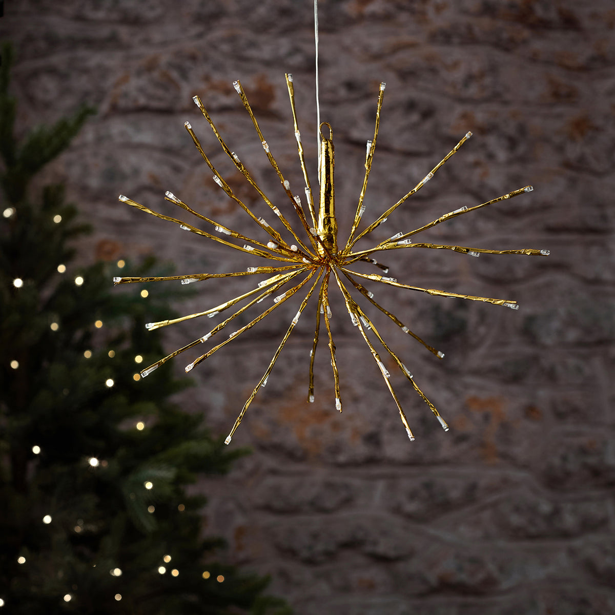 Detailaufnahme einer hängenden LED-Stern-Dekofigur mit warmweißer Beleuchtung in strahlenförmigem Design, positioniert vor grüner Baum-Silhouette und natürlichem Außenhintergrund – hochwertige Hänge-Lichterkette für gehobene Außen-Weihnachtsarrangements und elegante Fassaden-Dekorationen.