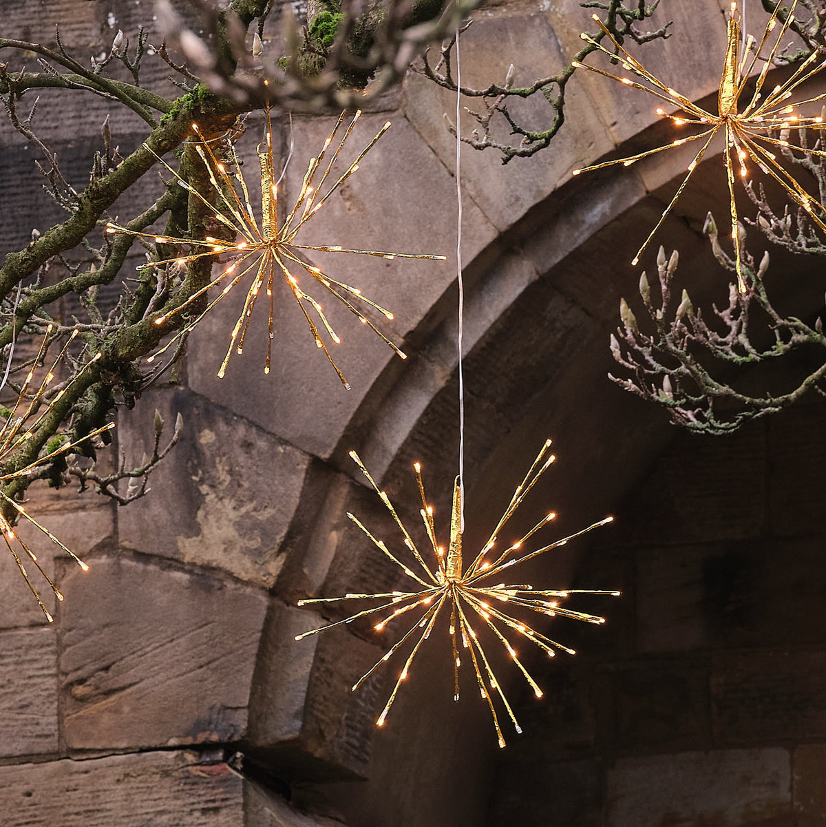 Charmante Anordnung von mehreren hängenden LED-Stern-Dekofiguren mit warmweißer Beleuchtung in strahlenförmigem Feuerwerk-Stil, positioniert an Gartenbogen und rustikalen Strukturen – dekorative Hänge-Lichterketten-Serie für umfassende Außen-Weihnachtsarrangements und gehobene Fassaden-Dekorationen.