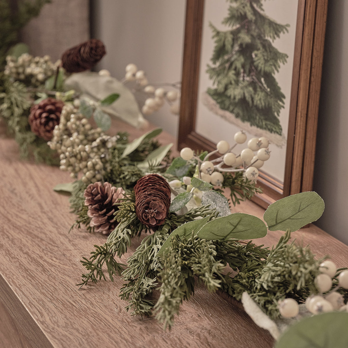 Charmantes üppiges Fensterbrett-Arrangement mit grünem Fichtengrün, burgundy strukturierten Blüten, weißen Beeren und natürlichen Holzakzenten, positioniert auf hellbraunem Holztisch vor Fensterrahmen mit Tannenbaum-Kunstdruck – festliche Weihnachts-Tischdekoration für Fensterdekorationen und elegante Wohnzimmer-Displays mit natürlichem Waldcharme.