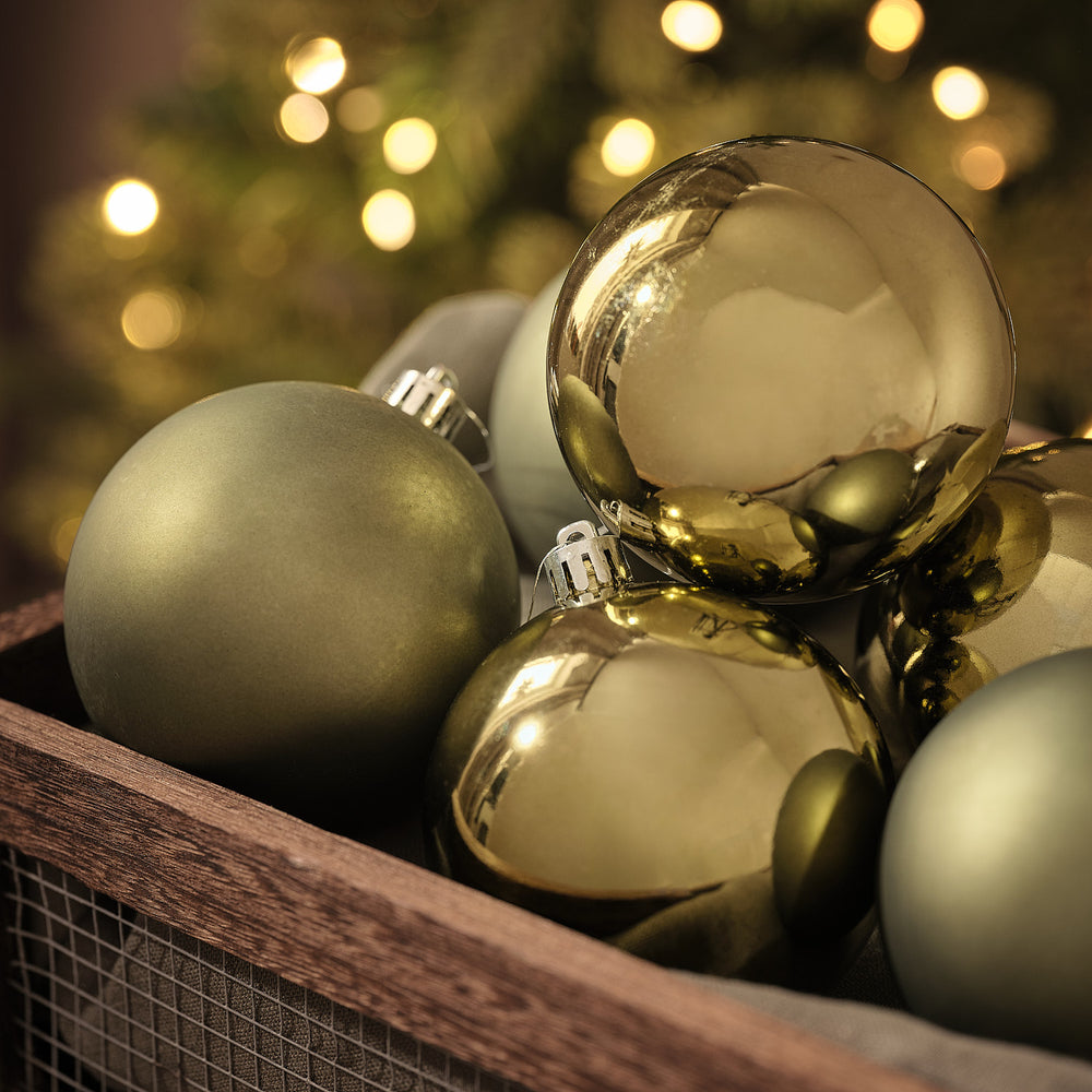 Goldene und olivgrüne Weihnachtskugeln in matter und glänzender Optik in Holzkiste vor beleuchtetem Tannenbaum.