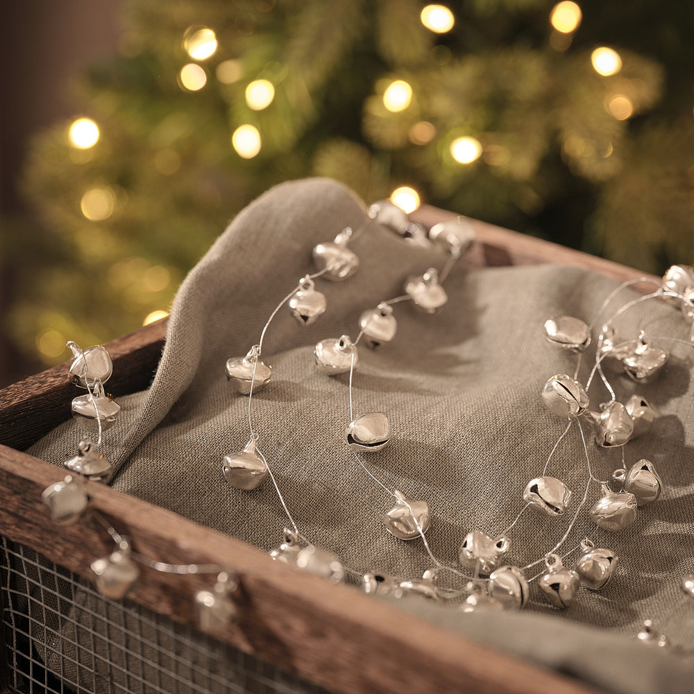 Perlenlichterkette mit Glöckchen auf Leinenstoff in Holzkiste vor Lichterglanz.