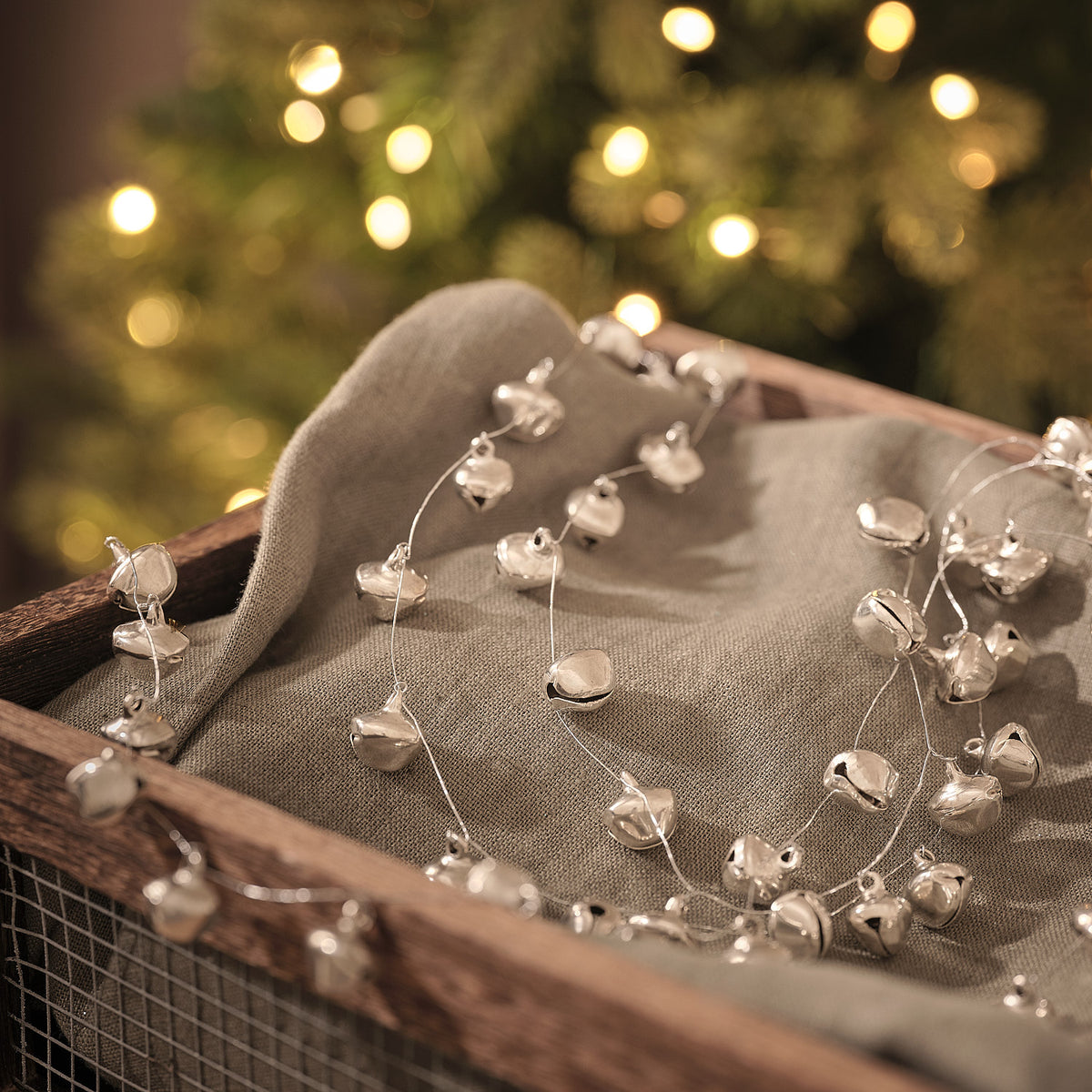 Perlenlichterkette mit Glöckchen auf Leinenstoff in Holzkiste vor Lichterglanz.