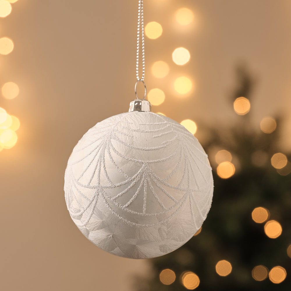 Weiße Weihnachtskugel mit silbernem Eiskristallmuster, hängend vor beleuchtetem Tannenbaum.