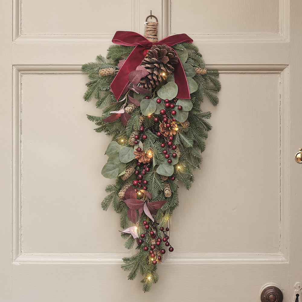 60 cm langer LED-Weihnachtszweig mit roten Beeren, Zapfen, grünen Blättern und Samtschleife an einer hellen Tür