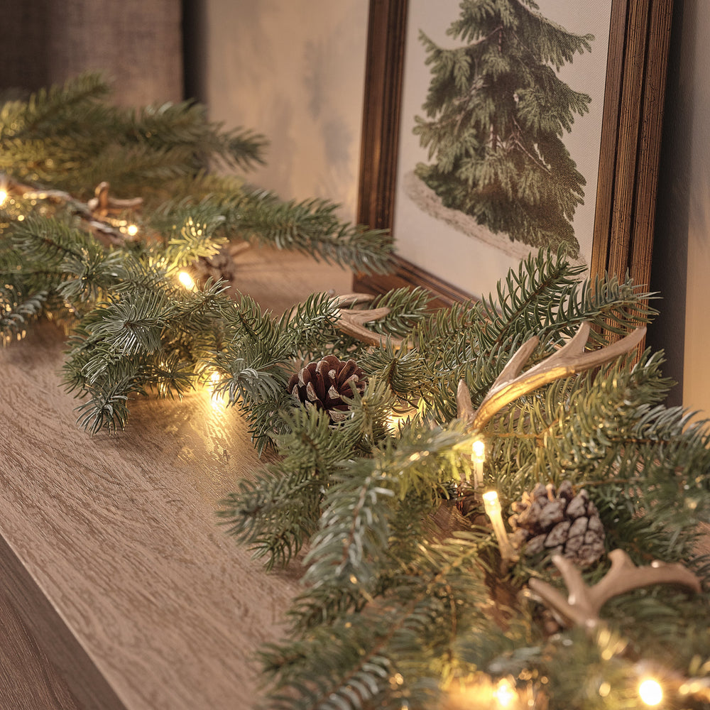 LED-Weihnachtsgirlande mit Geweih und Tannenzapfen auf Holzfläche vor gerahmtem Bild
