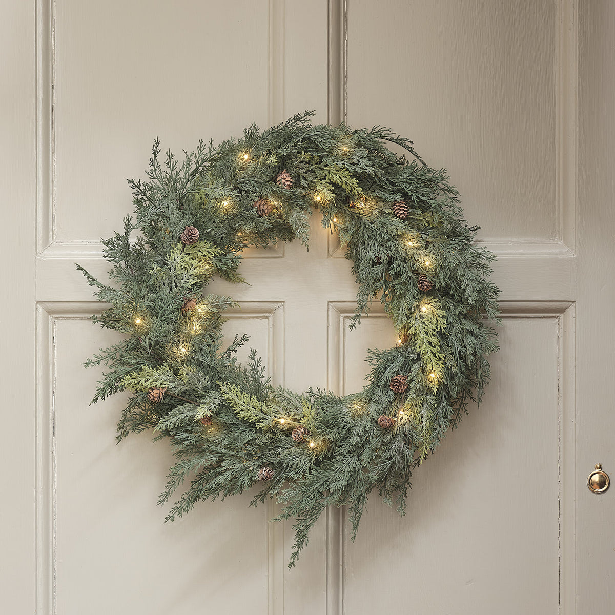 Weihnachtskranz mit Tannenzapfen und warmweißen LEDs an weißer Tür hängend