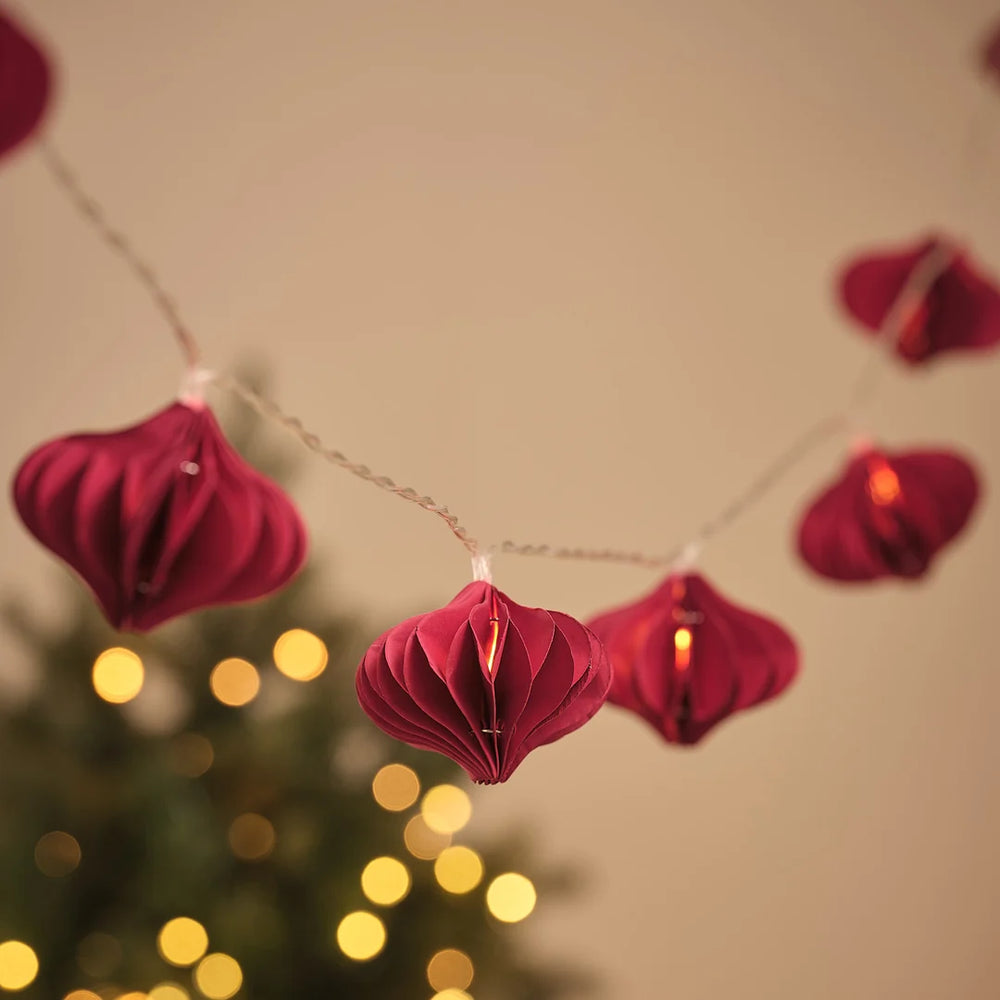 Lichterkette mit roten Papier-Weihnachtsanhängern vor Tannenbaum und Lichterglanz.