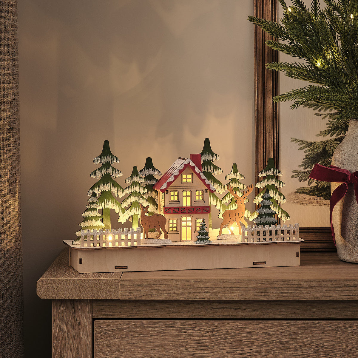 Beleuchtetes Holzhaus mit Rentieren und schneebedeckten Tannen auf einem Sideboard bei Dunkelheit