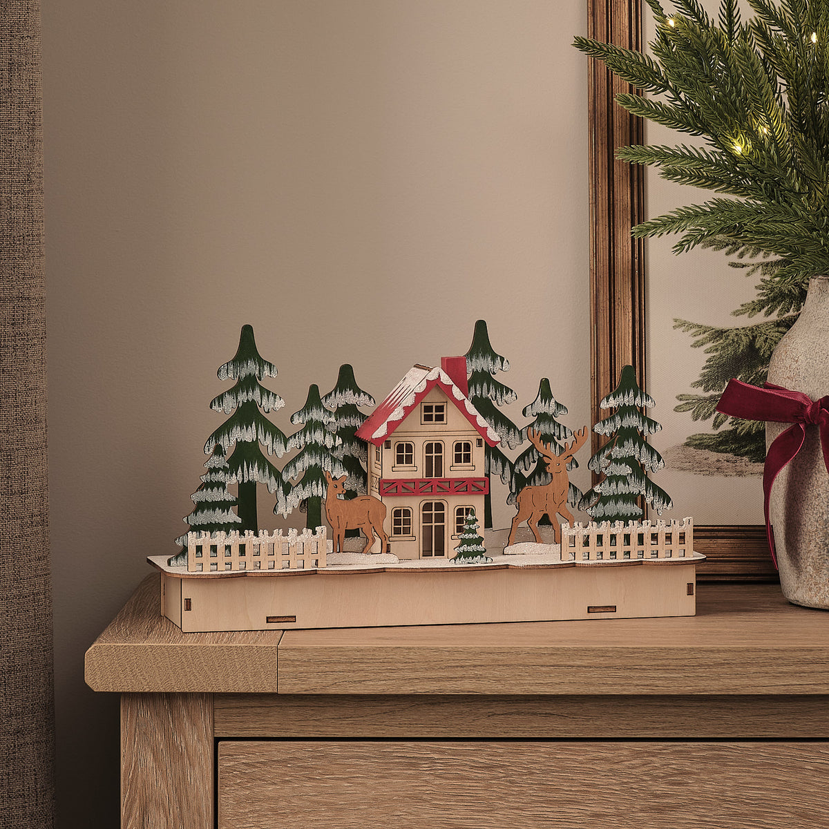 Unbeleuchtetes Holzhaus mit Rentieren und schneebedeckten Tannen auf einem Sideboard vor Wandbild