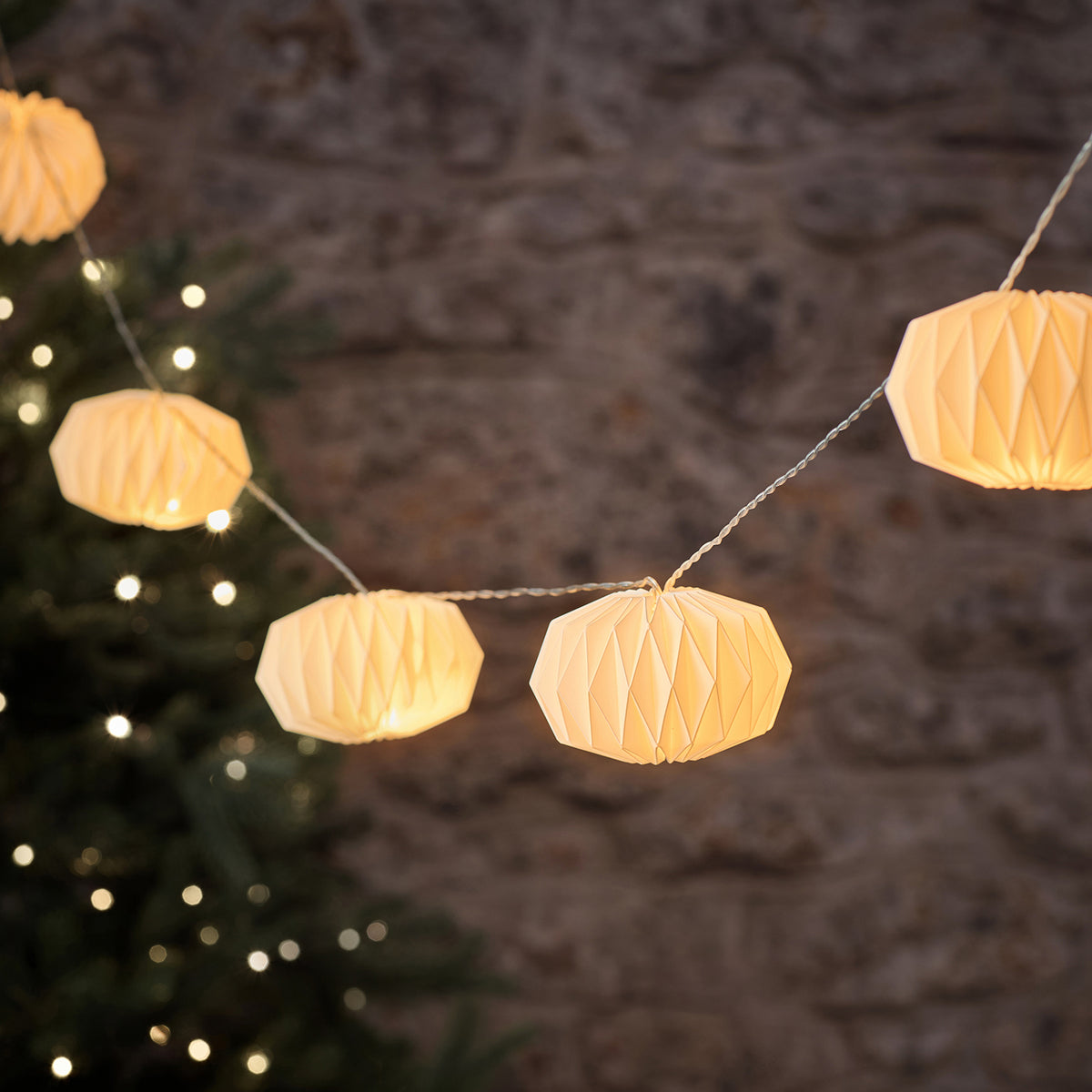 LED-Lichterkette mit mehreren weißen Papierlampions vor einer Steinwand und Tannenzweig
