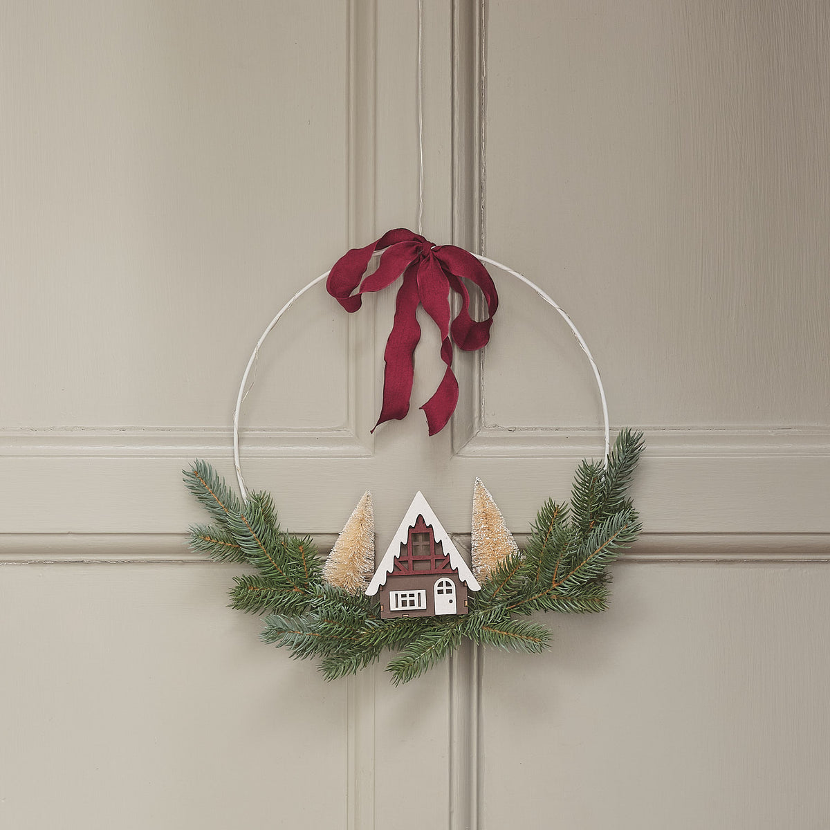LED-Weihnachtskranz mit Häuserszene aus Holz, Tannengrün und bordeauxroter Schleife ohne Beleuchtung an Wand