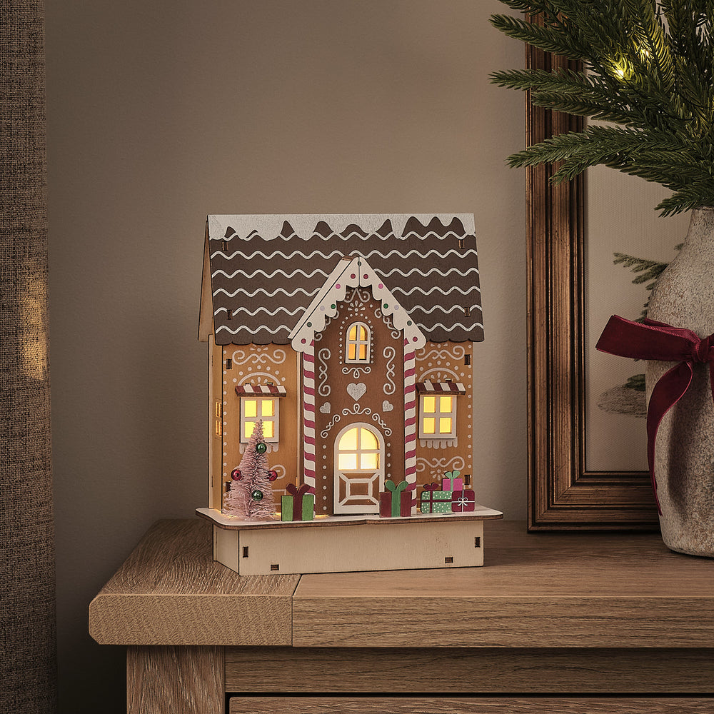 Beleuchtetes LED-Lebkuchenhaus aus Holz mit warmweißem Licht auf einem Holztisch neben einem Bilderrahmen