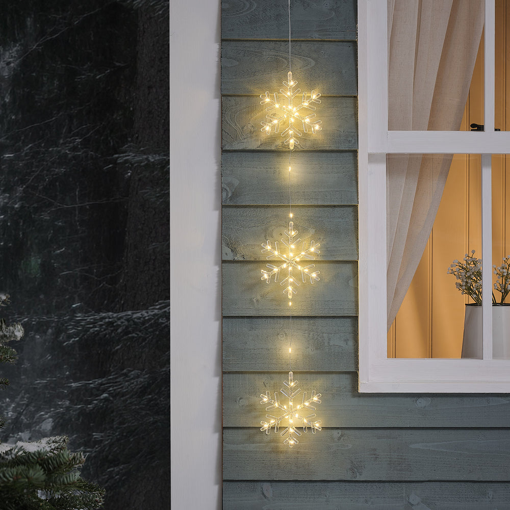 Drei warmweiß leuchtende LED-Schneeflocken hängen an einer Holzfassade neben einem Fenster