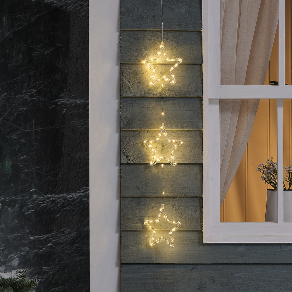 Drei leuchtende LED-Acrylsterne in warmweiß hängen vertikal an einer Außenwand neben einem Fenster.
