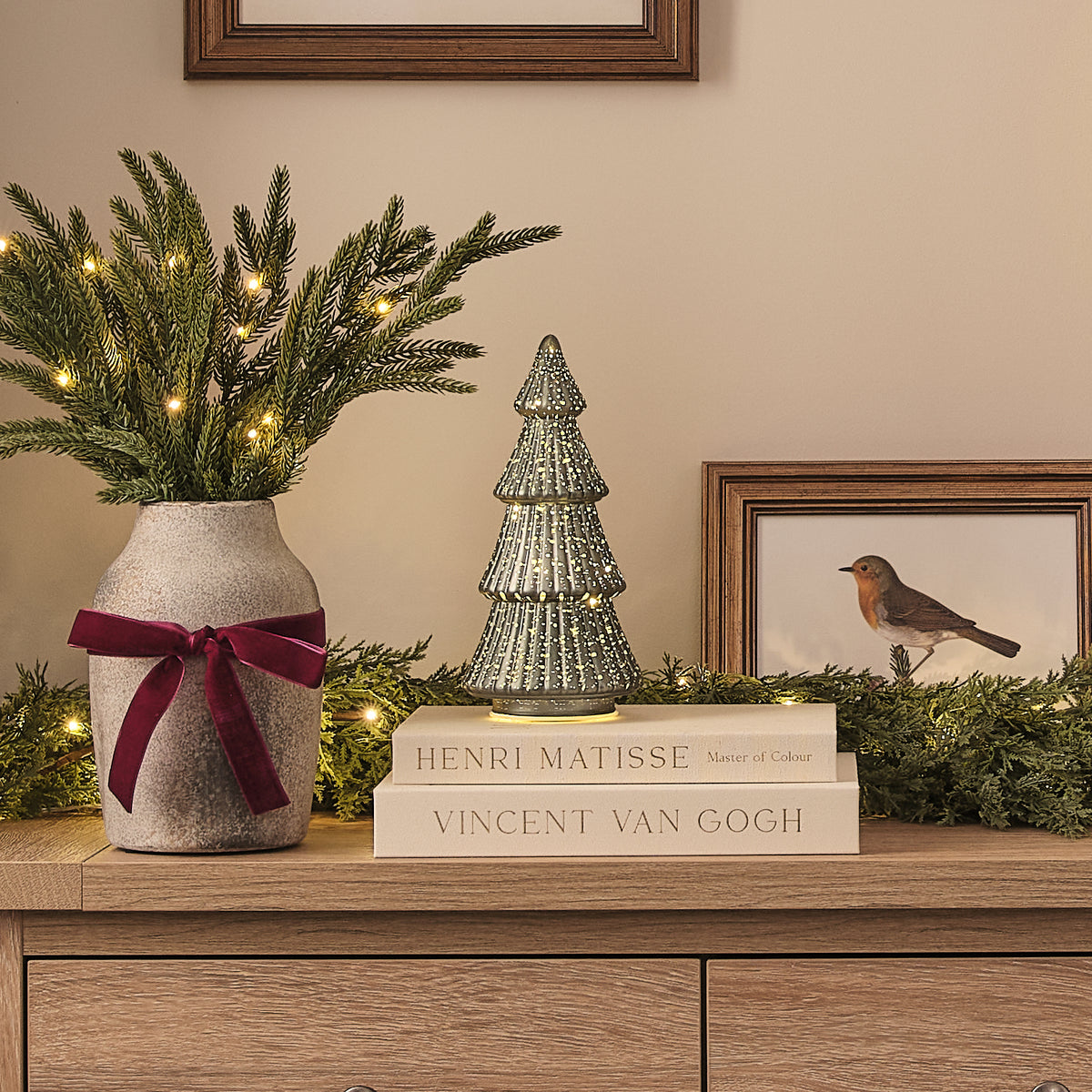 Grüner LED Tannenbaum aus Glas auf Büchern neben Vase mit Tannenzweigen und Lichterkette