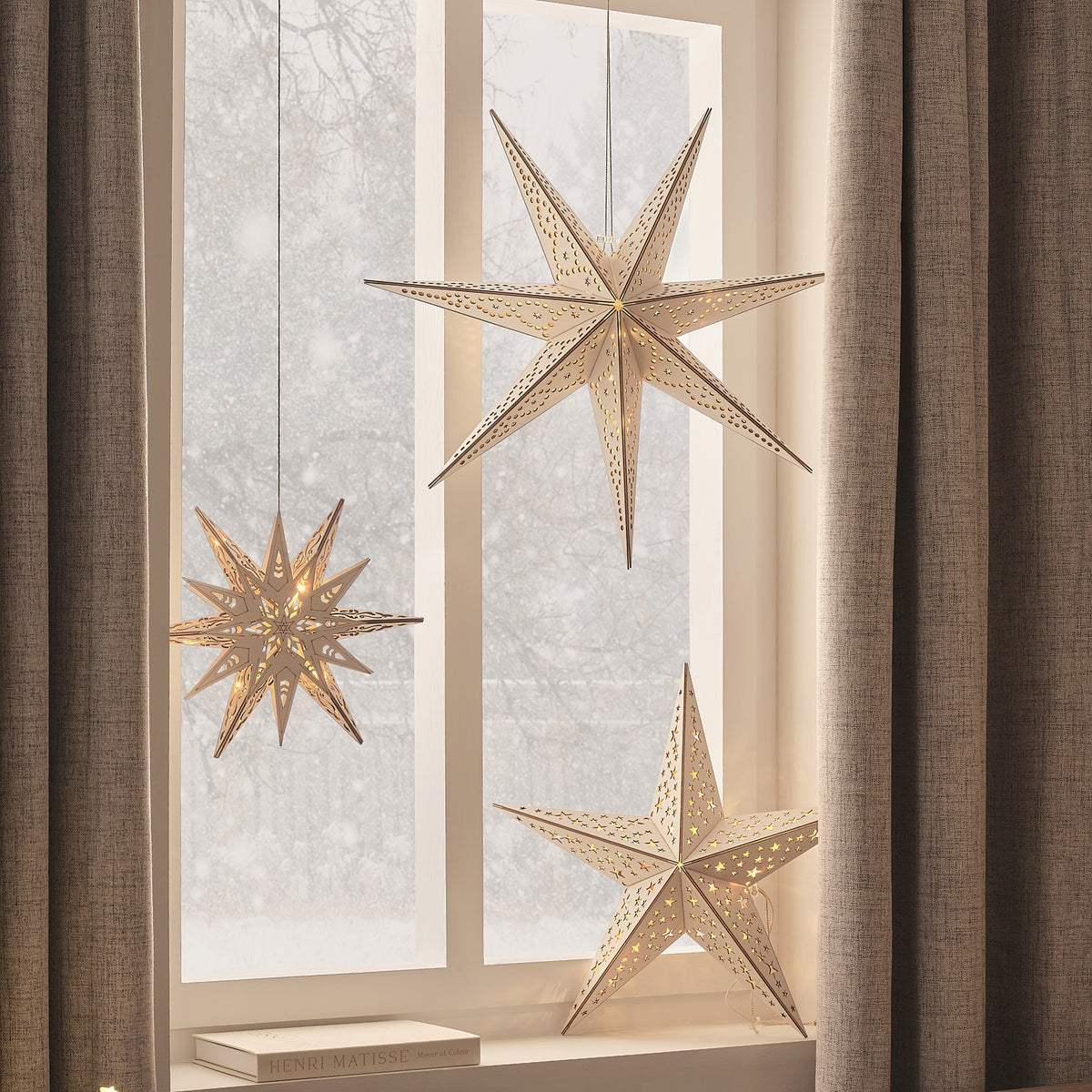 Drei unterschiedlich große LED-Holzsterne in Gold leuchten vor einem Fenster mit Vorhängen
