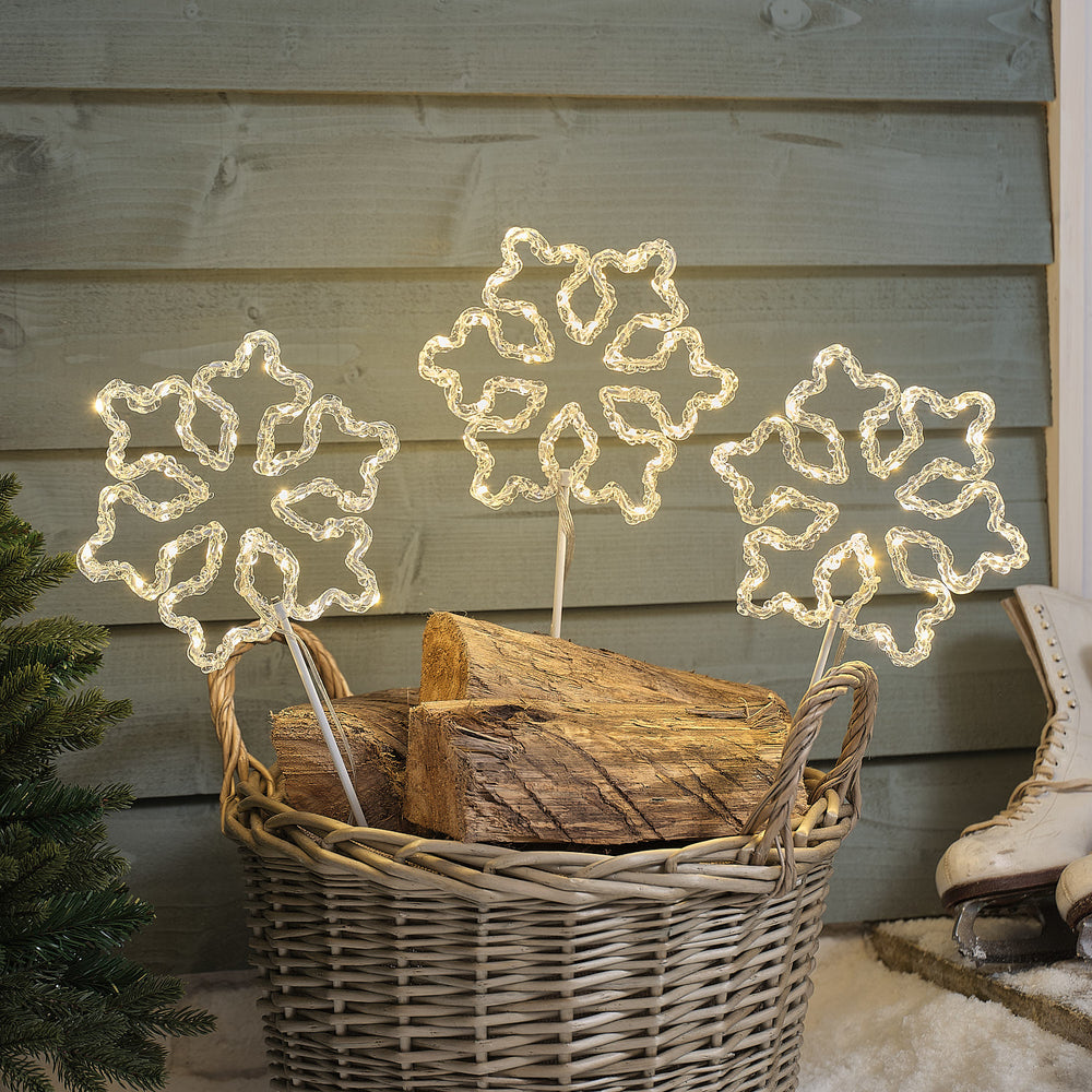 Drei beleuchtete Acryl-Schneeflocken Gartenstecker mit warmweißen LEDs in einem Korb mit Holzscheiten