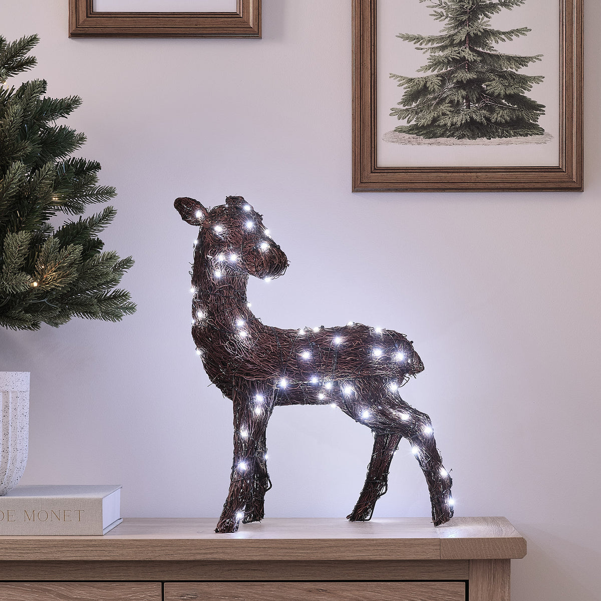 LED-Rentier aus Zweigen mit weißem Licht auf Sideboard vor Wand mit Bildern und Weihnachtsbaum