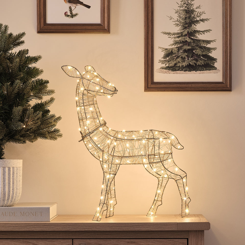 Goldenes LED-Rentier Hirschkuh mit warmweißen Mikro-LEDs auf Holzkommode neben Tannenzweig
