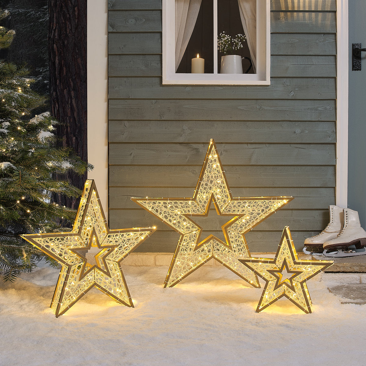 Drei beleuchtete goldene Sterne mit warmweißen LEDs auf Schnee vor Hauswand mit Fenster