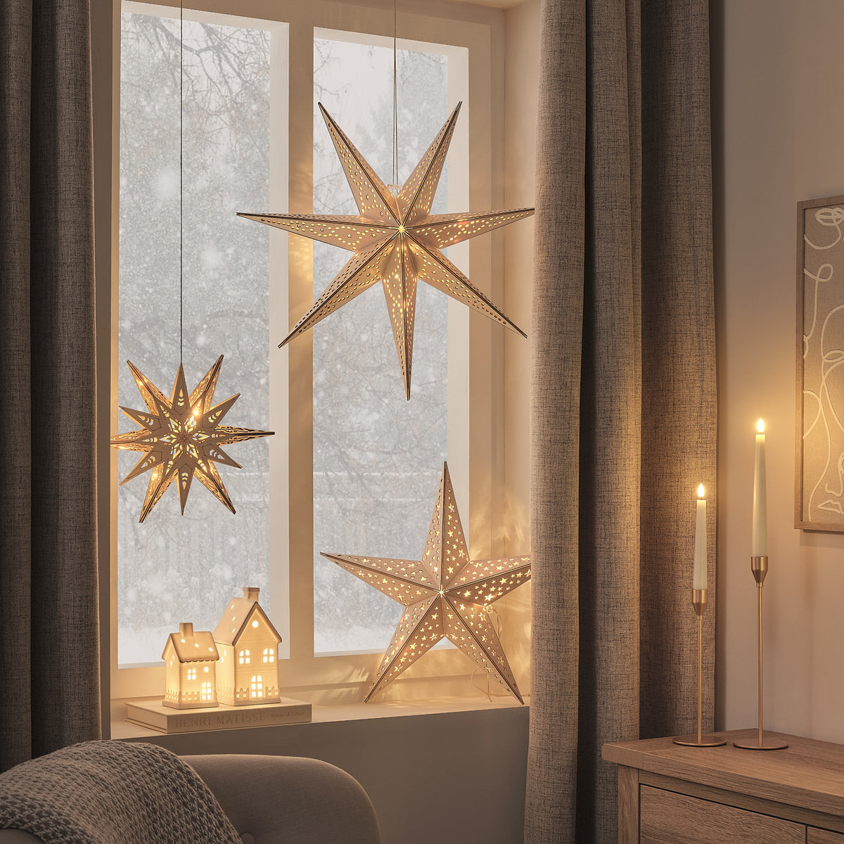 Wohnzimmerfenster mit drei beleuchteten Holzsternen und Kerzen auf Sideboard, warme Weihnachtsstimmung
