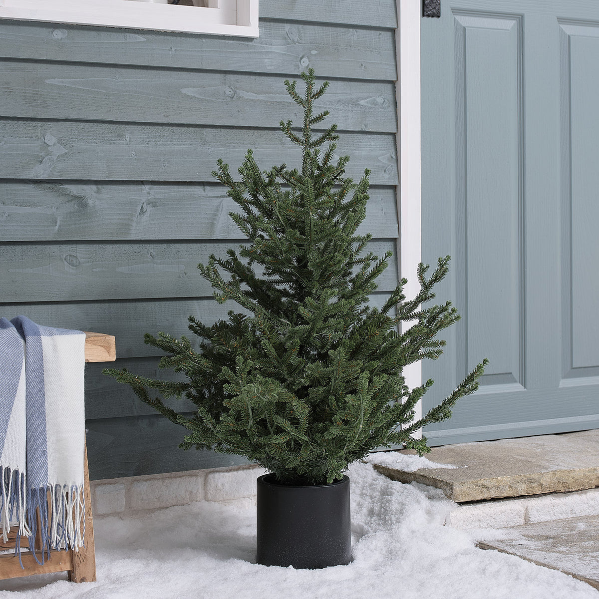 Beleuchteter künstlicher Weihnachtsbaum im schwarzen Topf vor Haustür auf Kunstschnee